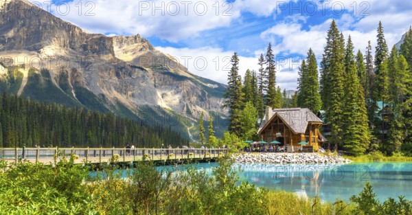 Wooden lodge sitting on the shore of a turquoise lake reflecting in the calm water with tourists walking on a bridge and a mountain in the background in yoho national park