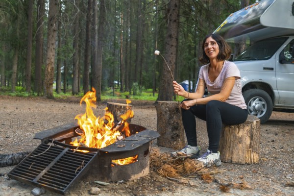 Female tourist roasting marshmallows over a campfire in a campground in banff national park, next to her camper van, enjoying the peace and quiet of nature
