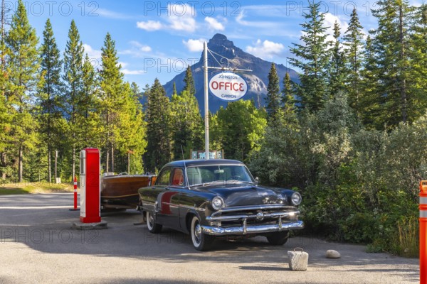 Vintage car towing a wooden boat trailer parked near a vintage gas pump by the johnston canyon office sign, with castle mountain and pine trees in the backdrop