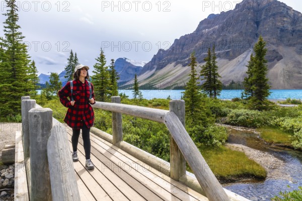 Female tourist with backpack is walking on wooden bridge and enjoying breathtaking view of turquoise bow lake and surrounding mountains in banff national park, canadian rockies, alberta, canada