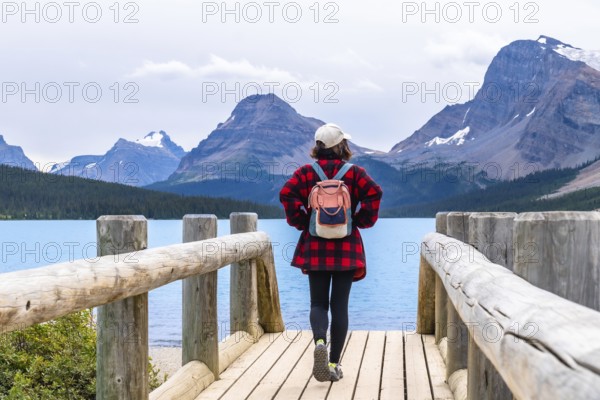 Female tourist with backpack walking on wooden bridge enjoying turquoise color water of bow lake and surrounding rocky mountains with glaciers and forests in banff national park, alberta, canada