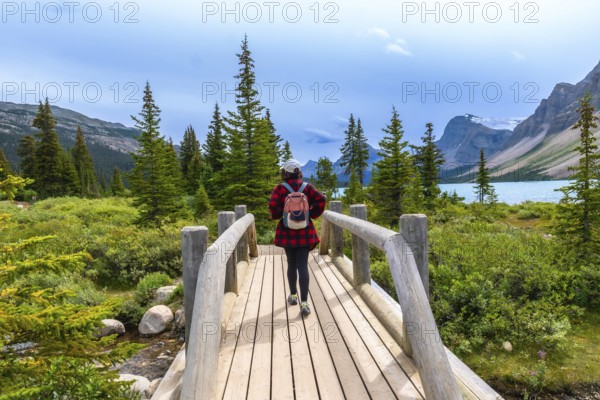 Female tourist with backpack walking on a wooden bridge at bow lake surrounded by lush vegetation and mountains. In banff national park. Canadian rockies. Alberta. Canada. During a cloudy summer day