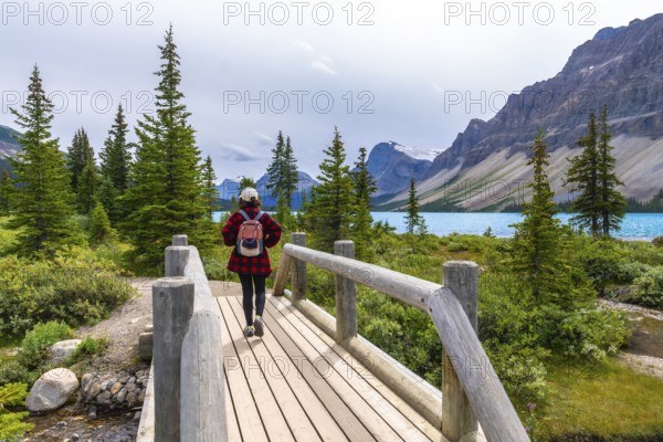 Female tourist walking on a wooden bridge and enjoying the breathtaking view of bow lake and surrounding mountains in banff national park, alberta, canada, during a cloudy summer day