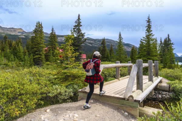Female tourist walking across a wooden bridge at bow lake, framed by a red roofed building, coniferous trees, and majestic mountains under a cloudy sky in banff national park