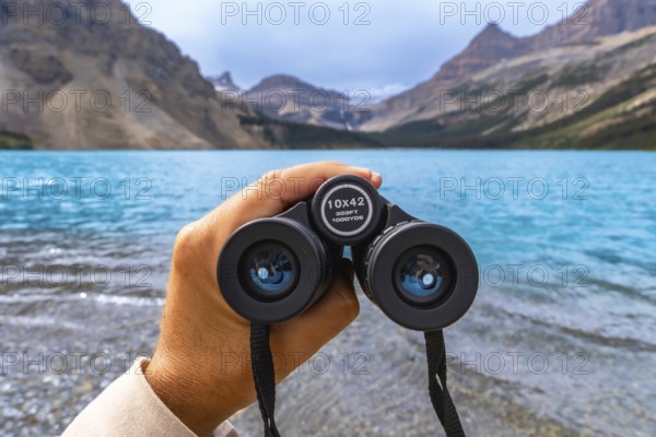 Hand holding binoculars overlooking the turquoise waters of bow lake with the canadian rockies in the background in banff national park, alberta, canada, during a sunny summer day