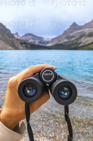 Hand holding binoculars, focusing on turquoise waters of bow lake surrounded by majestic mountains and glaciers in banff national park, alberta, canada
