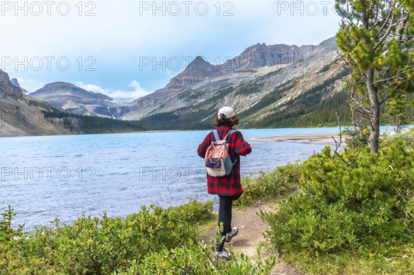 Female tourist with backpack walking along hiking trail by turquoise bow lake with crowfoot glacier and mountains in the background in banff national park, alberta, canada during a sunny summer day