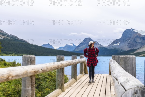 Female tourist walking along a wooden bridge and enjoying the breathtaking view of turquoise bow lake and surrounding mountains in banff national park, canadian rockies, alberta, canada