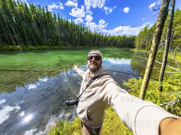 Photographer taking a selfie with outstretched arms at herbert lake, surrounded by turquoise water, lush forest, and a stunning sky, in banff national park, canadian rockies, alberta