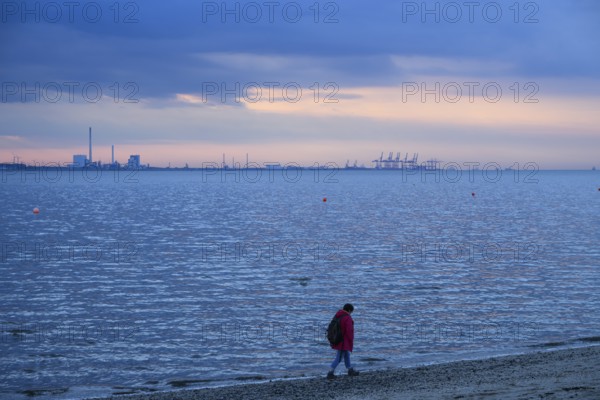 Person on the beach with a view of the sea and industrial areas in Wilhelmshafen right Jade-Weser Port, blue-orange twilight, Eckwarderhörne, Butjadingen, Lower Saxony, Germany