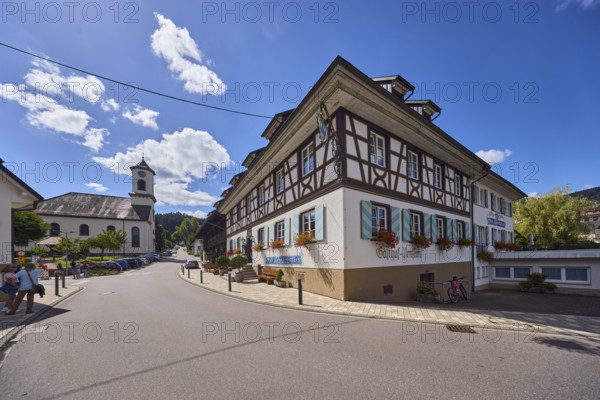Landgasthaus Drei Schneeballen, Gasthaus, St. Erhard church, historic building, house, sidewalk, street, barrier bollard, hill, coniferous forest, pedestrians as accessories, back light, side light, blue sky, cumulus clouds, intersection of main street with Friedhofstraße, Hofstetten, Black Forest, Ortenaukreis, Baden-Württemberg, Germany