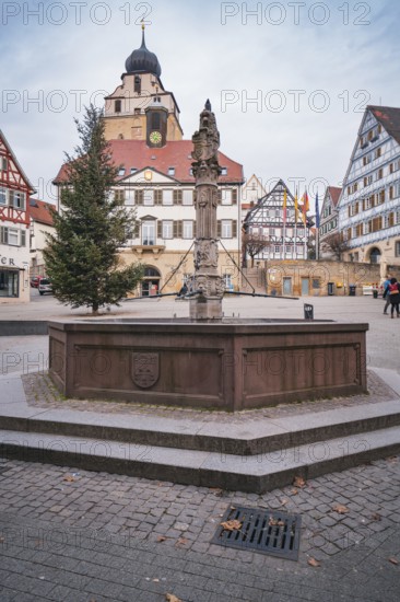 A fountain on a market square surrounded by historic buildings and a distinctive church tower under a cloudy sky, Herrenberg, Böblingen district, Germany