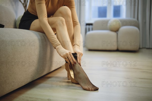Unrecognizable woman is seated on a couch in a bright room. She is putting on black fishnet stockings while dressed in a fitted top and a skirt. Natural light fills the space through large windows