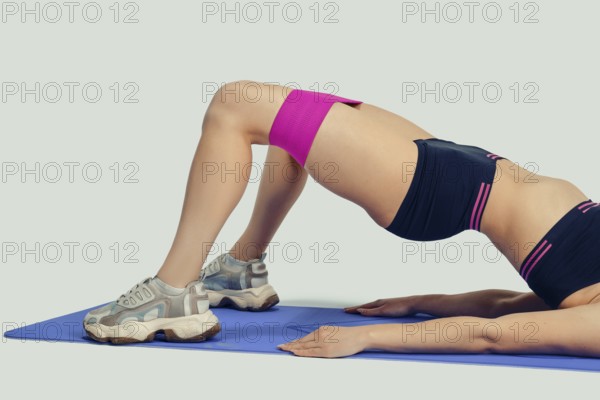 Unrecognizable woman lies on her back on a blue exercise mat, lifting her hips while using a resistance band on her thighs. She is in a home workout space