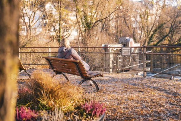 Woman sitting on park bench looking at river and prohibition sign in autumn, Stadtgarten Pforzheim, Germany