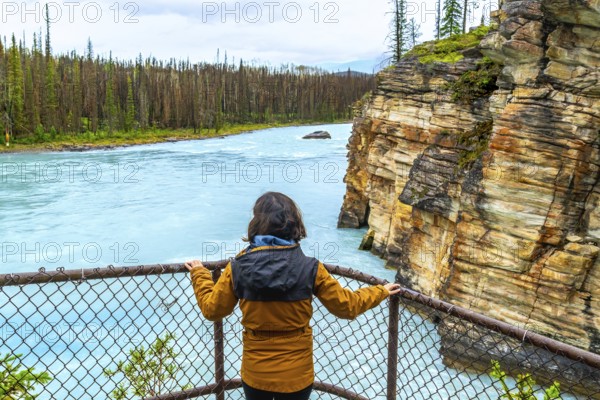 Tourist in a yellow jacket enjoying the breathtaking view of athabasca falls, surrounded by powerful turquoise waters, stunning rock formations, and lush pine forests