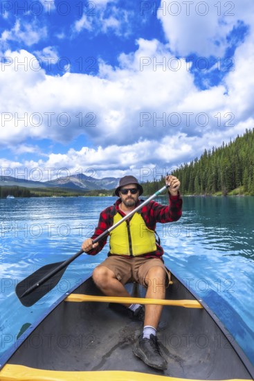 Tourist wearing sunglasses and a life jacket paddling a canoe on turquoise maligne lake under a bright blue sky, surrounded by lush forests and mountains in jasper national park, alberta
