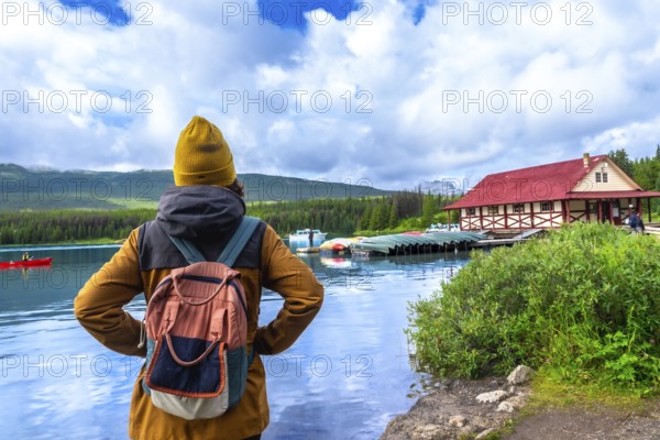 Female tourist wearing a backpack standing on the shore of maligne lake, admiring the iconic boathouse and canoes on a beautiful sunny day in jasper national park, alberta, canada