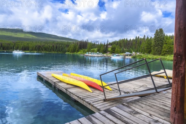 Colorful kayaks lined up on a wooden dock at maligne lake in jasper national park, alberta, while tourist boats navigate turquoise waters beneath a cloudy sky