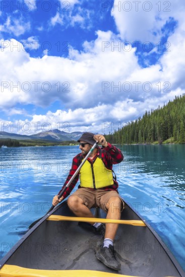 Tourist wearing sunglasses and life jacket paddling a canoe on beautiful maligne lake in jasper national park with blue sky, clouds, mountains, and forest in the background