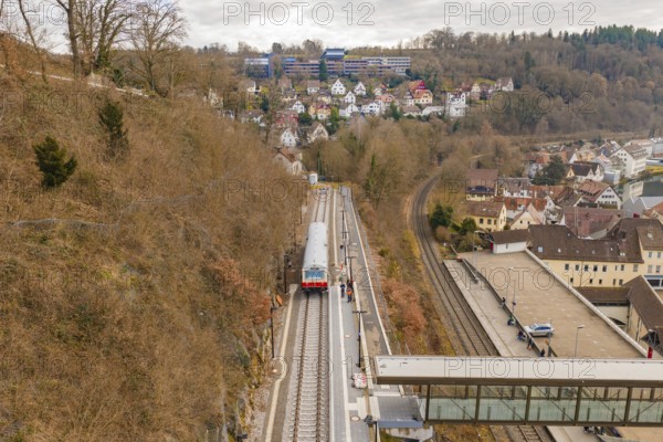 A train travels through a hilly urban landscape with wooded slopes and isolated houses under cloudy skies, historical event, first test run with a passenger train on the Hermann tracks, Hesse Railway since 1988, Calw, Calw district, Germany