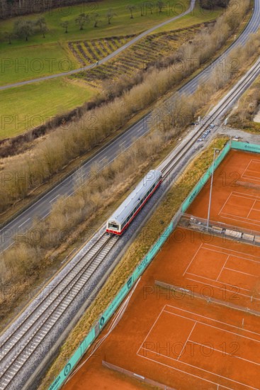 A train travels through rural landscape next to orange tennis courts on railroad tracks, historic event, first test run with a passenger train on the Hermann tracks, Hesse Railway since 1988, Calw district, Germany