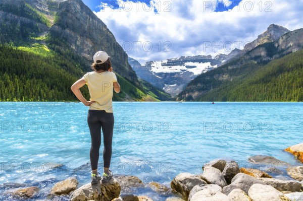 Tourist standing on rocks at the edge of vibrant turquoise waters of lake louise, admiring majestic mountains and glaciers in banff national park on a beautiful summer day