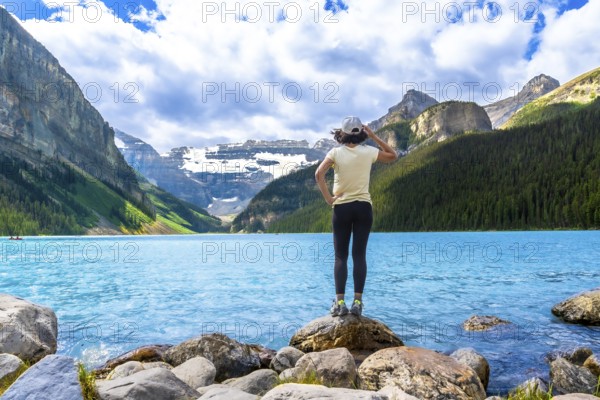 Tourist standing on rocks by the turquoise water of lake louise admires the surrounding canadian rockies in banff national park, alberta, on a sunny day with some clouds