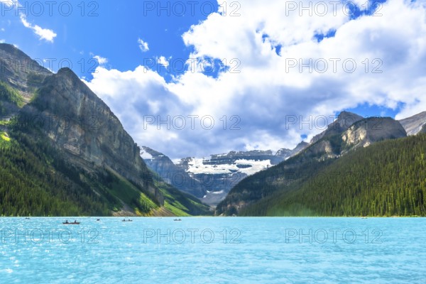 Canoes are paddling on a beautiful turquoise lake with surrounding mountains and glaciers in banff national park, alberta, canada, on a sunny summer day with white clouds and blue sky