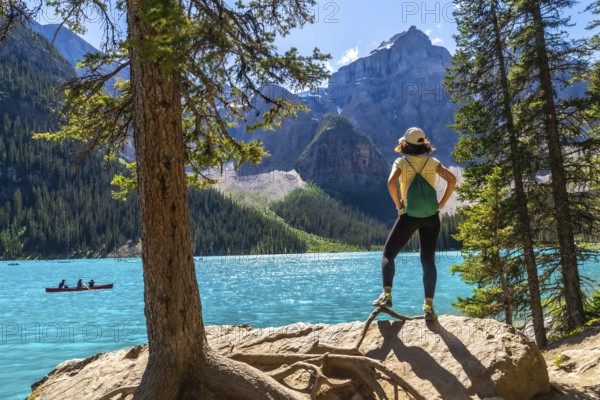 Female tourist standing on a rock by a tree, admiring the turquoise waters of moraine lake with canoeists and the majestic canadian rockies in the background