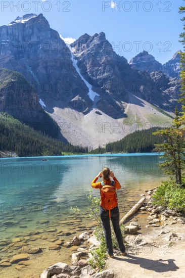 Female tourist with backpack taking pictures of the turquoise waters of moraine lake and the surrounding canadian rockies on a sunny summer day in banff national park, alberta, canada
