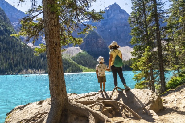 Mother and son holding hands on a rock by a tree, admiring the turquoise waters of moraine lake, surrounded by majestic mountains and lush pine forests in banff national park, alberta