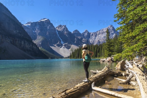 Female tourist standing on a log admiring the turquoise waters of moraine lake with the valley of the ten peaks in the background, banff national park, alberta, canada
