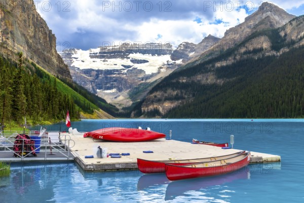 Red canoes are tied to a dock on the turquoise water of lake louise with a backdrop of snow capped mountains and evergreen forest in banff national park, alberta, canada