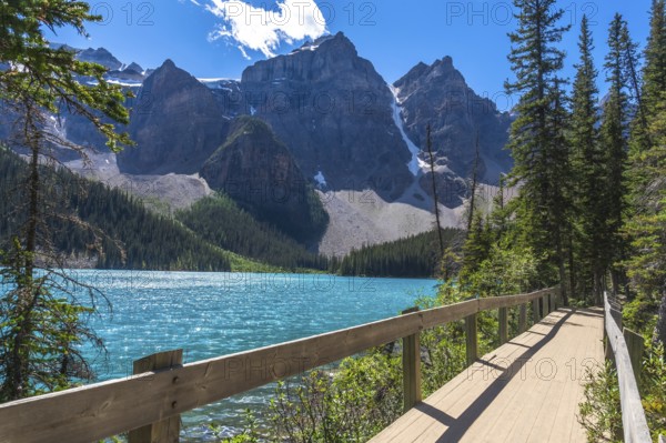 Moraine lake in banff national park features turquoise water, a wooden boardwalk, snow capped mountains, and lush vegetation during a sunny summer day in the canadian rockies