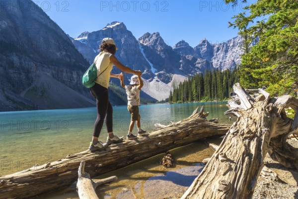 Mother and child balancing on a weathered log by the vibrant turquoise moraine lake, surrounded by the majestic peaks of the canadian rockies on a sunny summer day