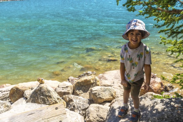 Happy child wearing a bucket hat standing by the turquoise water of moraine lake with a chipmunk eating on a rock in banff national park. Canadian rockies. Alberta. Canada. During a sunny summer day