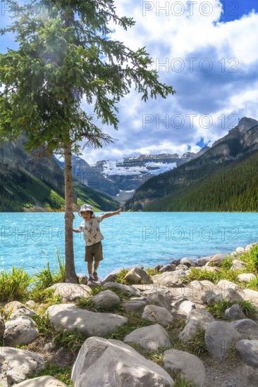 Young tourist balancing on rocks by a tree near the turquoise waters of lake louise, with snow capped mountains and a cloudy sky in the background, in banff national park, alberta, canada