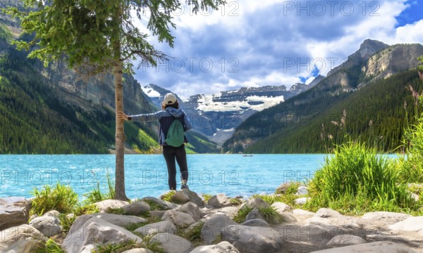 Female tourist standing by a tree on the rocky shore of turquoise lake louise, admiring the stunning view of surrounding mountains and glaciers in banff national park