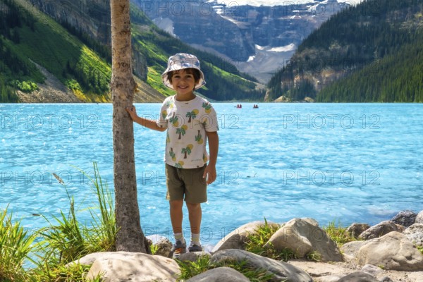 Young child standing by a tree on the rocky shore of turquoise lake louise, taking in the breathtaking scenery of banff national park with distant kayakers