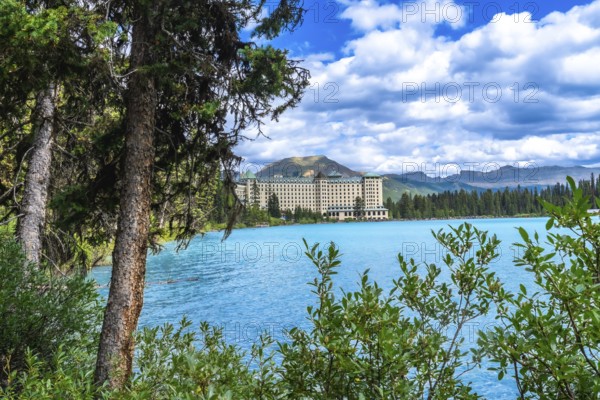 Lush vegetation framing turquoise waters of lake louise reflects a luxury hotel against stunning mountain peaks and a cloudy summer sky in banff national park, alberta