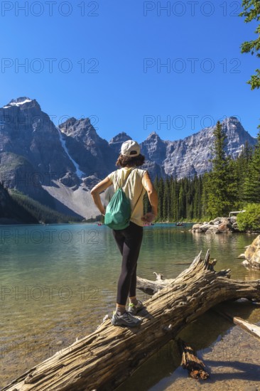 Female tourist standing on a log, admiring the turquoise waters of moraine lake, surrounded by the majestic canadian rockies, in banff national park, alberta, canada, on a sunny summer day