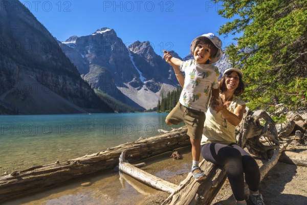 Mother and son are having fun while posing on a log by the turquoise waters of moraine lake, surrounded by the majestic mountains of banff national park on a sunny day