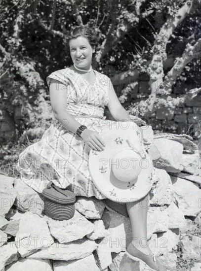 Portrait of young woman wearing summer dress sitting on stone wall holding straw hat, Malta, Europe, 1956