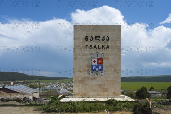 A monument with the coat of arms of Tsalka against a village backdrop and a blue sky, town sign Tsalka, Kartli, Georgia
