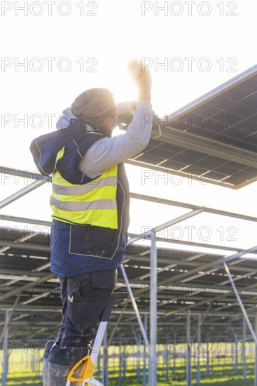 Worker mounts solar panel on scaffolding, stands on ladder with yellow safety vest, energy transition, construction of PV open space, Baden-Württemberg, Germany