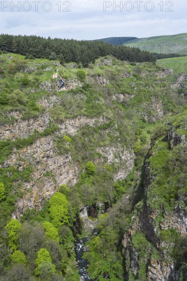 A green gorge with rocks and trees crossed by a small river under a cloudy sky, man riding a bicycle on a stretched rope, Dashbashi Gorge, Khrami River, Chrami, Tsalka Nature Reserve, Kartli, Georgia
