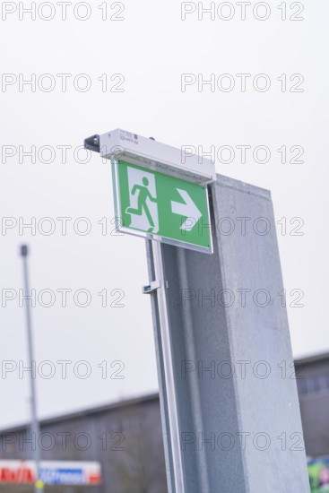 Green emergency exit sign with directional arrow on a metal post, parking garage construction with charging infrastructure, Calw, Germany