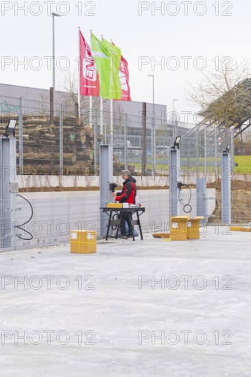 A worker sitting at a table next to a fence with assembled cables and boxes in an industrial area, parking garage construction with charging infrastructure, Calw, Germany