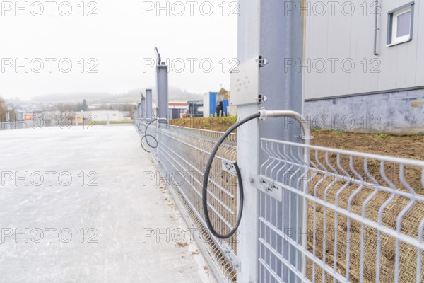 A cable is attached to a fence along a concrete surface. Industrial environment in the background, car park construction with charging infrastructure, Calw, Germany